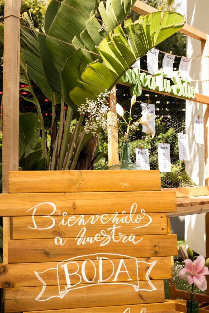 Cartel de madera de bienvenida a la boda junto al seating plan, entre plantas tropicales