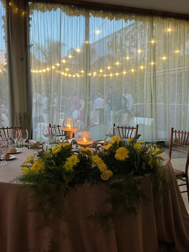 Mesa con flores amarillas y velas frente a cortinas con guirnalda de luces