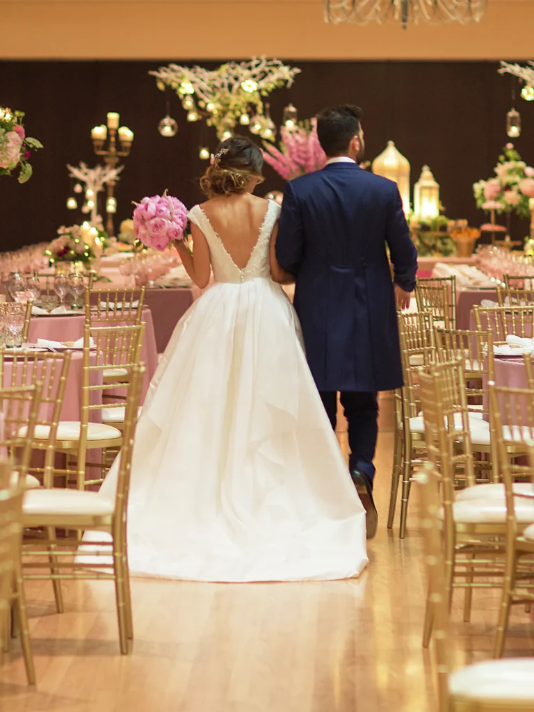 Novios caminando por el pasillo del salón de banquetes, entre mesas con flores rosas