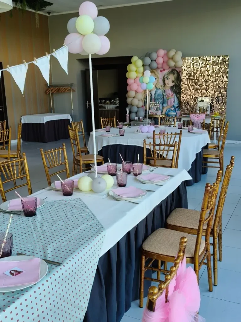 Mesas de una comunión con temática Top Model en el salón, con globos pastel y la mesa de dulces al fondo